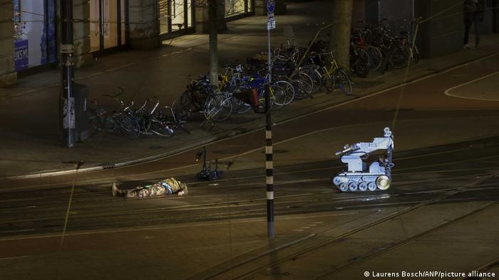 A man in camouflage is lying next to a police robot on the street