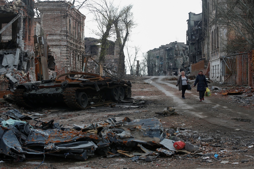 People walking near a destroyed tank and destroyed buildings in the context of the Ukraine-Russia conflict.